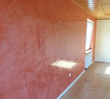 Heller, frisch renovierter Raum mit rötlich strukturierter Akzentwand, Holzdecke, Fischgrat‑Parkett und großem Fenster mit einfallendem Tageslicht